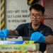 A young man wearing blue nitrile gloves and a dark polo shirt with a visible arm tattoo is handling food service materials near a 'Stretch-Seal' food wrap box. He is working in what appears to be an industrial or military vessel environment, indicated by the safety signage and metal grating in the background. The setting suggests a food service or galley operation.