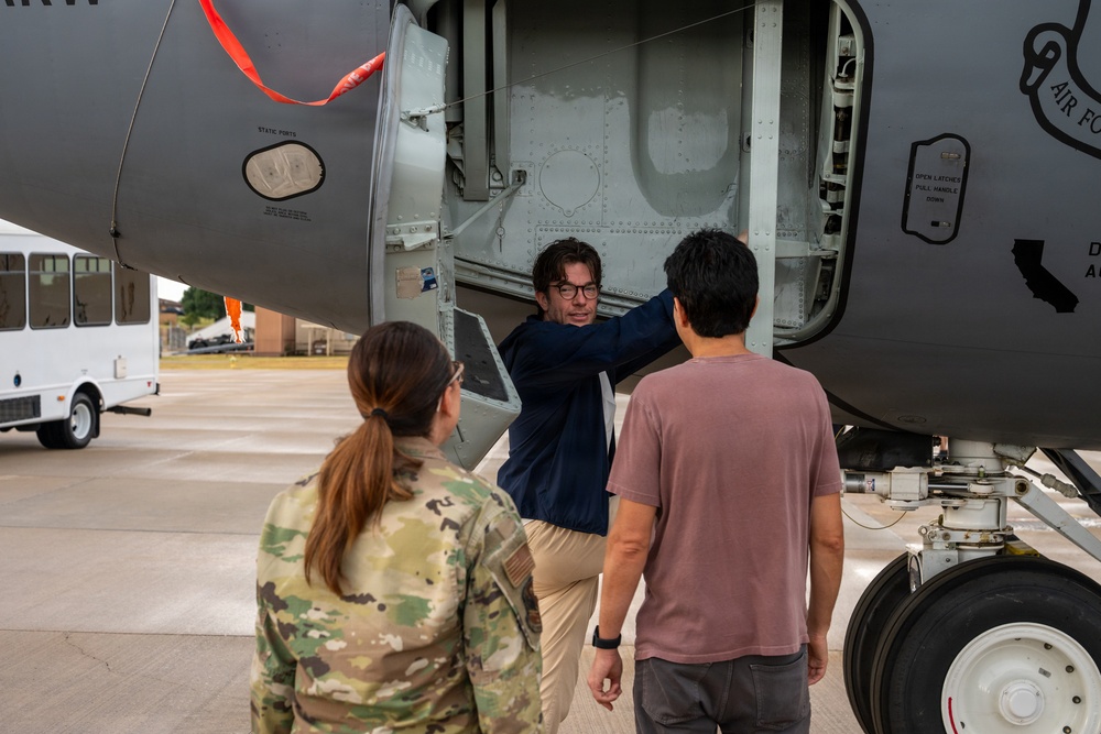 Three individuals are gathered near the underside of a large military aircraft, examining an open access panel or equipment bay. One person in a navy blue shirt and glasses is pointing into the open compartment, while a uniformed military personnel in camouflage and a civilian in a pink shirt observe. The setting appears to be a military airfield or tarmac.