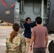 Three individuals are gathered near the underside of a large military aircraft, examining an open access panel or equipment bay. One person in a navy blue shirt and glasses is pointing into the open compartment, while a uniformed military personnel in camouflage and a civilian in a pink shirt observe. The setting appears to be a military airfield or tarmac.