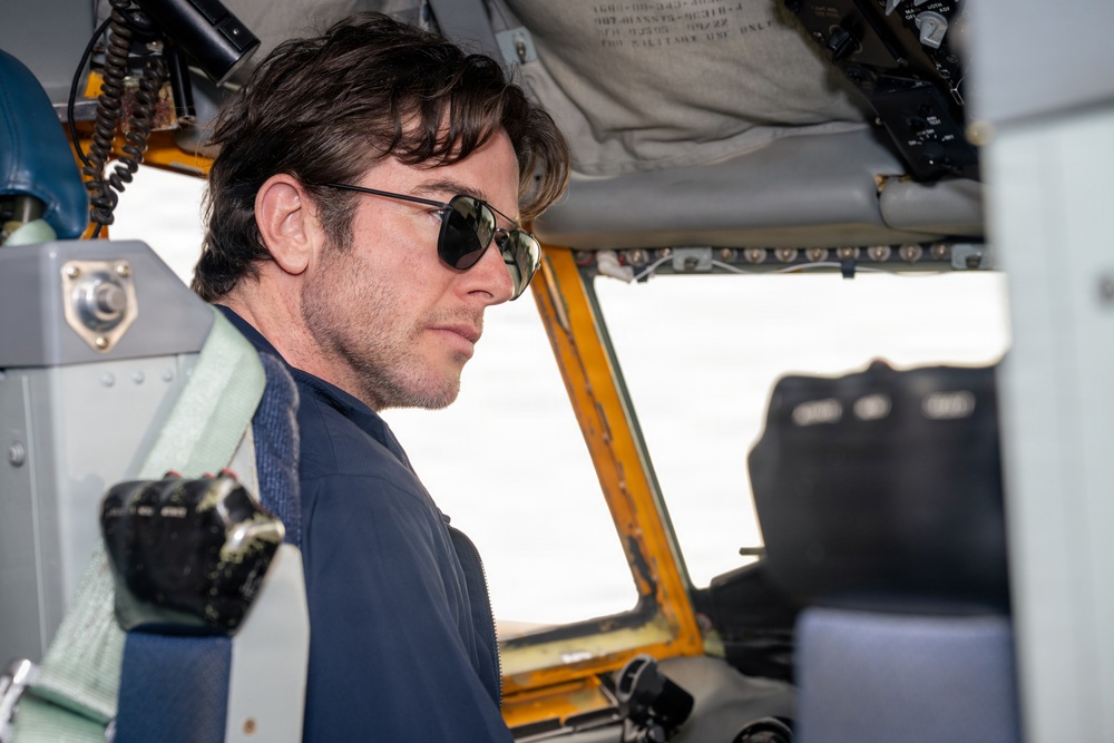 A man with dark hair and aviator-style sunglasses is seated inside what appears to be a vintage or military aircraft cockpit. He is wearing a dark zip-up jacket and a harness/seatbelt, gazing forward through the windscreen. The cockpit interior shows aged yellow framing, various instruments, and overhead equipment.
