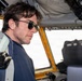 A man with dark hair and aviator-style sunglasses is seated inside what appears to be a vintage or military aircraft cockpit. He is wearing a dark zip-up jacket and a harness/seatbelt, gazing forward through the windscreen. The cockpit interior shows aged yellow framing, various instruments, and overhead equipment.