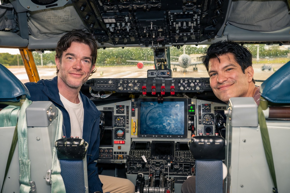 Two men are seated in the cockpit of a large aircraft, posing and smiling for the camera. The cockpit features an extensive array of instruments, controls, switches, and a central display screen. The windshield shows a wet, rain-speckled view of an airport tarmac with other aircraft visible in the background.