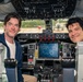 Two men are seated in the cockpit of a large aircraft, posing and smiling for the camera. The cockpit features an extensive array of instruments, controls, switches, and a central display screen. The windshield shows a wet, rain-speckled view of an airport tarmac with other aircraft visible in the background.