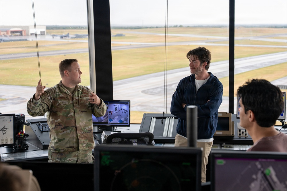A military service member in camouflage uniform appears to be briefing or explaining something to two civilians inside what appears to be an air traffic control tower. The setting overlooks an airfield with runways and aircraft visible through large floor-to-ceiling windows. Multiple monitoring screens and control equipment are visible throughout the workspace.
