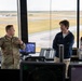 A military service member in camouflage uniform appears to be briefing or explaining something to two civilians inside what appears to be an air traffic control tower. The setting overlooks an airfield with runways and aircraft visible through large floor-to-ceiling windows. Multiple monitoring screens and control equipment are visible throughout the workspace.