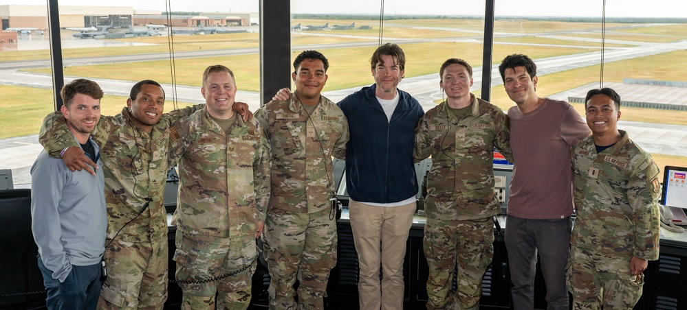 A group of eight individuals pose for a photo inside what appears to be a military air traffic control tower. Six of the individuals are wearing U.S. Air Force camouflage uniforms (OCP pattern), while two are in civilian clothing. An active airfield with aircraft and runways is visible through the large windows behind them.