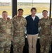 A group of eight individuals pose for a photo inside what appears to be a military air traffic control tower. Six of the individuals are wearing U.S. Air Force camouflage uniforms (OCP pattern), while two are in civilian clothing. An active airfield with aircraft and runways is visible through the large windows behind them.