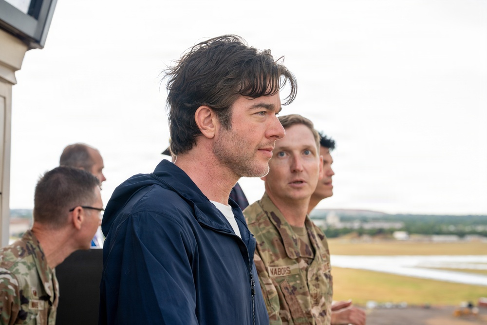 A group of individuals, including at least two military personnel in camouflage uniforms, stand on what appears to be an elevated observation platform overlooking an airfield or military installation. A civilian man in a navy blue jacket is prominently featured in the foreground, appearing to observe the area below. The setting suggests an official visit or briefing at a military air base.
