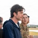 A group of individuals, including at least two military personnel in camouflage uniforms, stand on what appears to be an elevated observation platform overlooking an airfield or military installation. A civilian man in a navy blue jacket is prominently featured in the foreground, appearing to observe the area below. The setting suggests an official visit or briefing at a military air base.