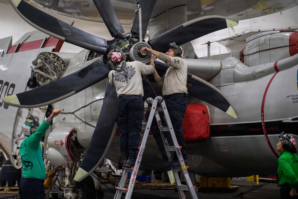 Military aviation mechanics from VAW-113 'Mech Shop' are performing maintenance on a large turboprop aircraft engine and propeller assembly in what appears to be a carrier hangar bay. Two technicians stand on a ladder working directly on the propeller hub while two others assist from ground level. The aircraft features a multi-blade propeller configuration consistent with a Northrop Grumman E-2 Hawkeye or C-2 Greyhound.