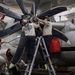 Military aviation mechanics from VAW-113 'Mech Shop' are performing maintenance on a large turboprop aircraft engine and propeller assembly in what appears to be a carrier hangar bay. Two technicians stand on a ladder working directly on the propeller hub while two others assist from ground level. The aircraft features a multi-blade propeller configuration consistent with a Northrop Grumman E-2 Hawkeye or C-2 Greyhound.