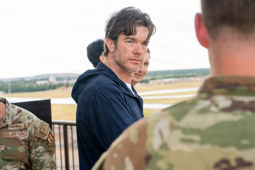 A civilian man in a navy blue hoodie jacket is standing on what appears to be an elevated observation area at a military airfield, surrounded by uniformed U.S. Air Force personnel. The man, who has dark wet hair and light stubble, appears to be looking back over his shoulder toward the camera. The setting suggests an official visit or tour of a military installation.