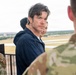 A civilian man in a navy blue hoodie jacket is standing on what appears to be an elevated observation area at a military airfield, surrounded by uniformed U.S. Air Force personnel. The man, who has dark wet hair and light stubble, appears to be looking back over his shoulder toward the camera. The setting suggests an official visit or tour of a military installation.
