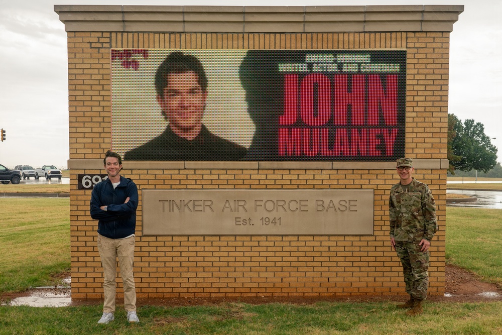 Two individuals stand in front of the entrance sign for Tinker Air Force Base, established in 1941. The base's LED marquee display shows a promotional image and text for 'John Mulaney,' described as an award-winning writer, actor, and comedian. One person is in civilian clothing and the other is in military camouflage uniform.