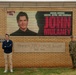 Two individuals stand in front of the entrance sign for Tinker Air Force Base, established in 1941. The base's LED marquee display shows a promotional image and text for 'John Mulaney,' described as an award-winning writer, actor, and comedian. One person is in civilian clothing and the other is in military camouflage uniform.