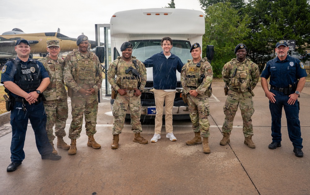 A group of eight individuals pose for a photo in a rainy outdoor setting, consisting of military personnel in camouflage uniforms, two civilian law enforcement officers in dark blue uniforms, and one civilian in casual clothing. They are standing in front of a white shuttle bus, with what appears to be a military aircraft static display visible in the background. The setting appears to be a military installation or air base.