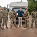 A group of eight individuals pose for a photo in a rainy outdoor setting, consisting of military personnel in camouflage uniforms, two civilian law enforcement officers in dark blue uniforms, and one civilian in casual clothing. They are standing in front of a white shuttle bus, with what appears to be a military aircraft static display visible in the background. The setting appears to be a military installation or air base.