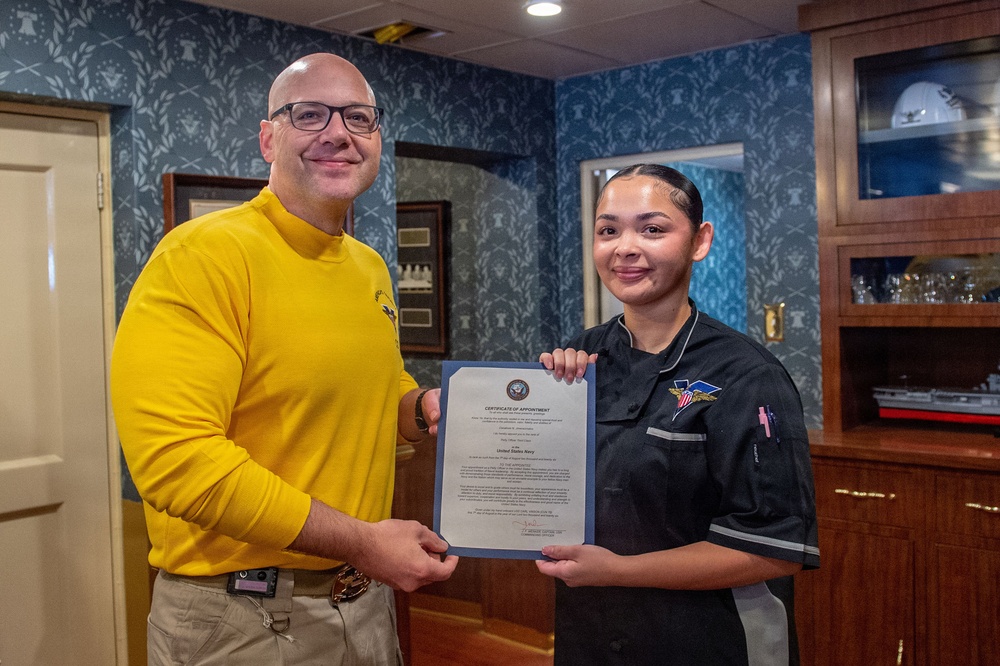 Two individuals are posing together while jointly holding a formal document that reads 'Certificate of Appointment' from the United States Navy. The man on the left is wearing a yellow long-sleeve shirt, while the younger woman on the right is wearing a black uniform with an embroidered patch. The setting appears to be an official Navy office or wardroom with decorative blue wallpaper and wooden cabinetry.
