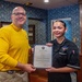 Two individuals are posing together while jointly holding a formal document that reads 'Certificate of Appointment' from the United States Navy. The man on the left is wearing a yellow long-sleeve shirt, while the younger woman on the right is wearing a black uniform with an embroidered patch. The setting appears to be an official Navy office or wardroom with decorative blue wallpaper and wooden cabinetry.