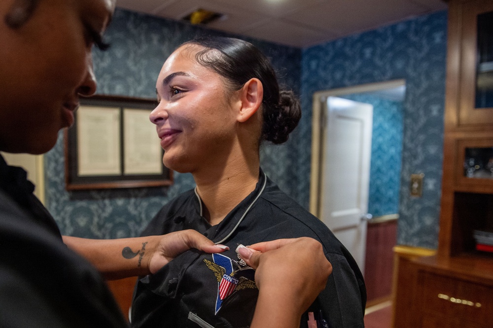A young woman in a black uniform is having a patch or pin affixed to her chest by another person. The woman receiving the insignia is smiling emotionally, suggesting this is a meaningful ceremonial moment such as a pinning ceremony. The setting appears to be a formal office or ceremonial room with decorative wallpaper and framed documents on the wall.
