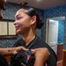 A young woman in a black uniform is having a patch or pin affixed to her chest by another person. The woman receiving the insignia is smiling emotionally, suggesting this is a meaningful ceremonial moment such as a pinning ceremony. The setting appears to be a formal office or ceremonial room with decorative wallpaper and framed documents on the wall.