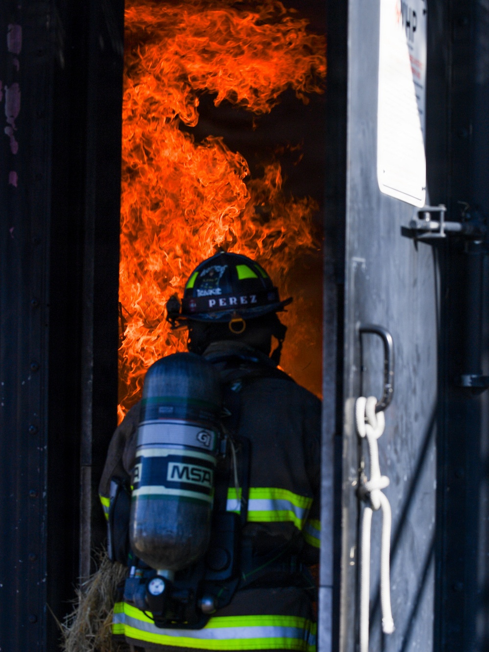 A firefighter identified as 'PEREZ' is positioned at the threshold of a doorway engulfed in intense orange flames. The firefighter is equipped with full turnout gear and an MSA self-contained breathing apparatus (SCBA). The image captures a high-risk moment of active firefighting operations, likely an interior attack or controlled burn training exercise.