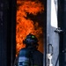 A firefighter identified as 'PEREZ' is positioned at the threshold of a doorway engulfed in intense orange flames. The firefighter is equipped with full turnout gear and an MSA self-contained breathing apparatus (SCBA). The image captures a high-risk moment of active firefighting operations, likely an interior attack or controlled burn training exercise.