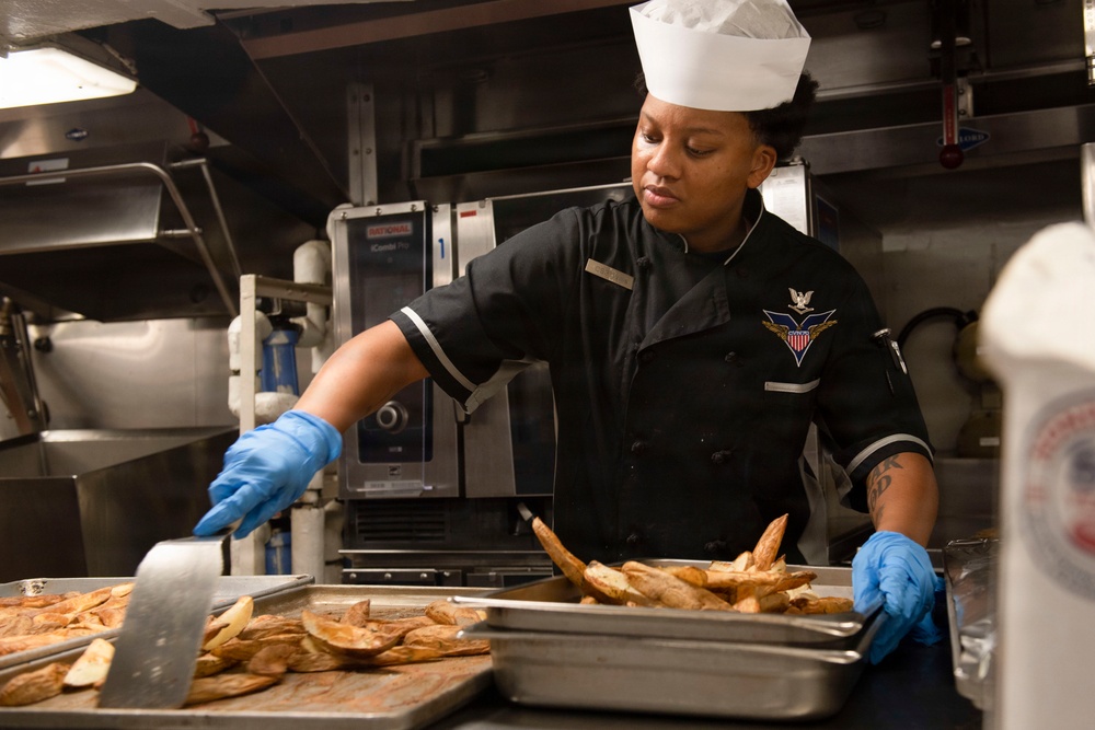 A U.S. Navy culinary specialist is working in a military galley kitchen, handling trays of roasted potato wedges. She is wearing a black Navy uniform with an insignia patch, a white chef's hat, and blue latex gloves. The setting appears to be an institutional or shipboard kitchen with commercial cooking equipment visible in the background.