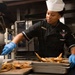 A U.S. Navy culinary specialist is working in a military galley kitchen, handling trays of roasted potato wedges. She is wearing a black Navy uniform with an insignia patch, a white chef's hat, and blue latex gloves. The setting appears to be an institutional or shipboard kitchen with commercial cooking equipment visible in the background.