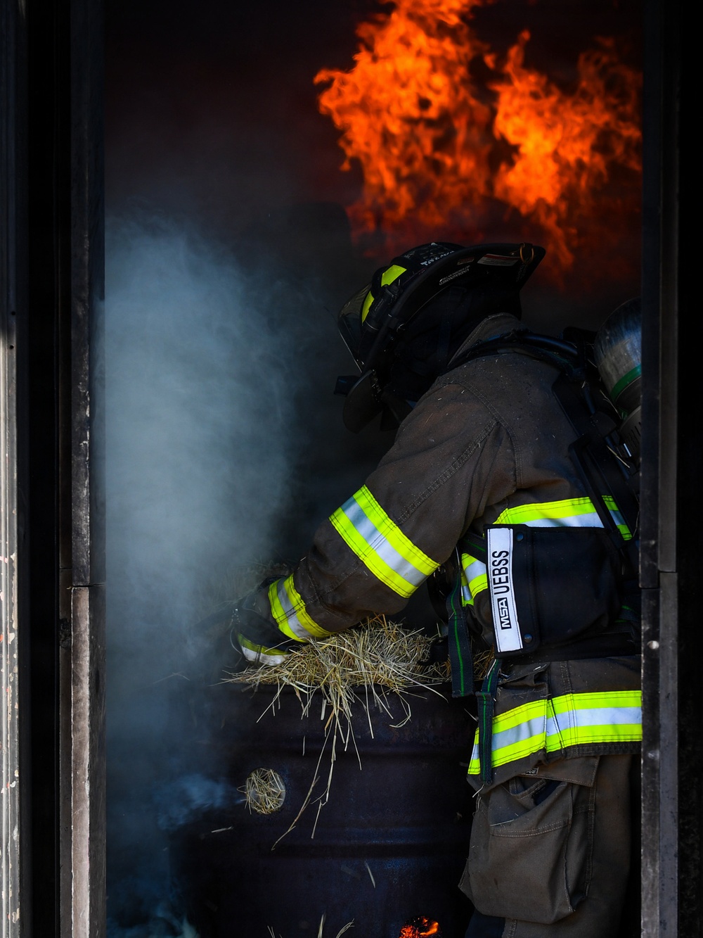A firefighter in full turnout gear is positioned in a doorway or opening engulfed in smoke and flames. The firefighter appears to be handling straw or hay, possibly as fuel material, suggesting this may be a controlled burn or training exercise. Large orange flames are visible in the background above the smoke-filled space.