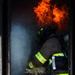A firefighter in full turnout gear is positioned in a doorway or opening engulfed in smoke and flames. The firefighter appears to be handling straw or hay, possibly as fuel material, suggesting this may be a controlled burn or training exercise. Large orange flames are visible in the background above the smoke-filled space.