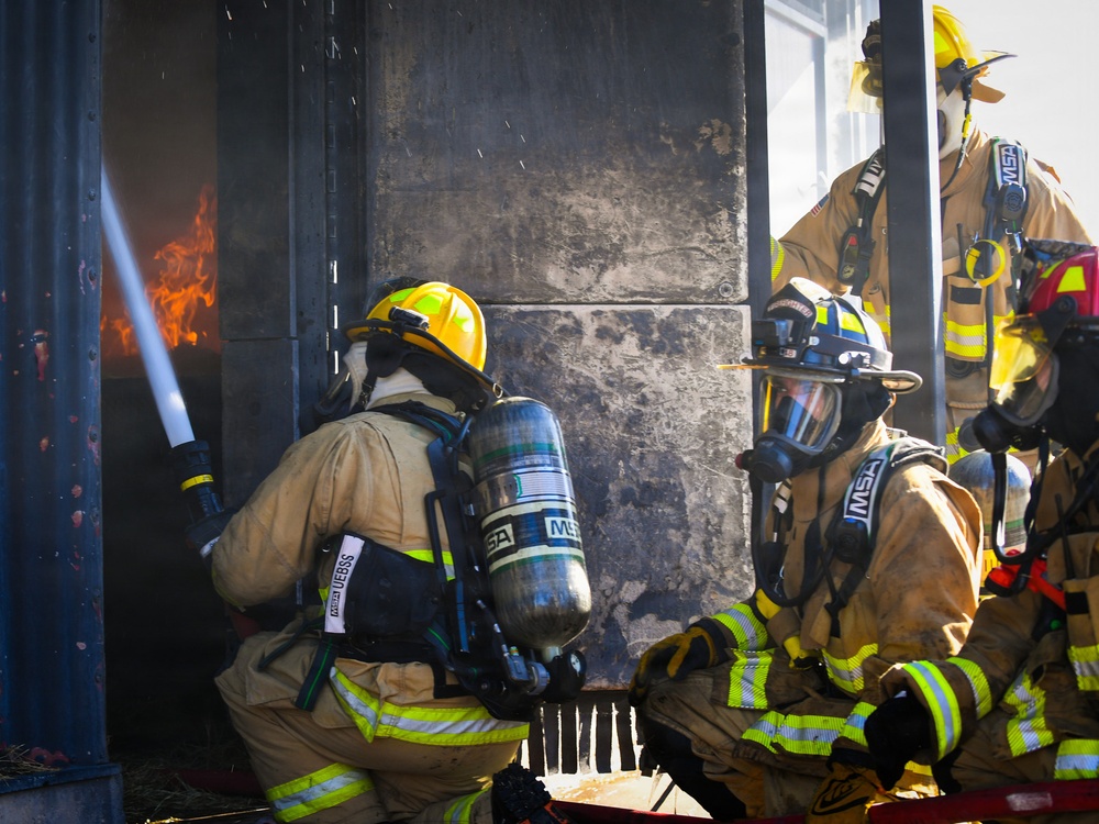 Multiple firefighters in full personal protective equipment are actively combating a fire using a hose line directed into a burning structure. The scene appears to be either a live fire training exercise or an active structural fire response, with visible flames and soot-blackened walls. At least three firefighters are visible in close proximity, wearing MSA-branded SCBA (Self-Contained Breathing Apparatus) units.