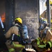 Multiple firefighters in full personal protective equipment are actively combating a fire using a hose line directed into a burning structure. The scene appears to be either a live fire training exercise or an active structural fire response, with visible flames and soot-blackened walls. At least three firefighters are visible in close proximity, wearing MSA-branded SCBA (Self-Contained Breathing Apparatus) units.