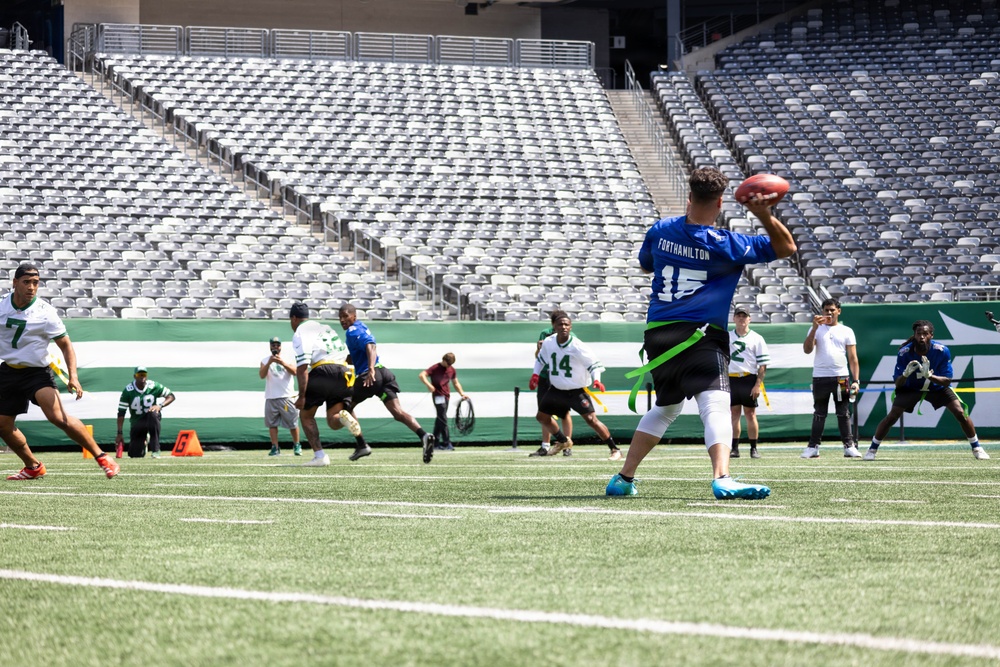 A flag football game is being played on a professional NFL stadium field, identifiable by the New York Jets branding on the end zone wall. The foreground player wearing a blue jersey numbered 15 with the name 'FORTHAMILTON' is in a throwing stance holding a football. The stadium seats are entirely empty, suggesting this is a community or youth event rather than a professional game.