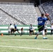 A flag football game is being played on a professional NFL stadium field, identifiable by the New York Jets branding on the end zone wall. The foreground player wearing a blue jersey numbered 15 with the name 'FORTHAMILTON' is in a throwing stance holding a football. The stadium seats are entirely empty, suggesting this is a community or youth event rather than a professional game.
