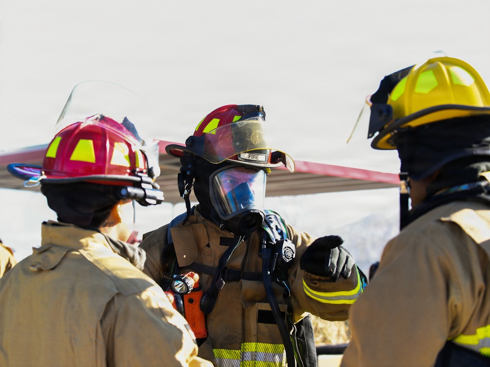 Three firefighters in full protective gear are gathered together, appearing to be in discussion or briefing. The central firefighter is wearing a full-face SCBA (Self-Contained Breathing Apparatus) mask and a red helmet labeled 'FIRE', while the others wear red and yellow helmets respectively. The setting appears to be outdoors in a bright, overcast environment, possibly at a training exercise or incident scene.