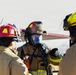 Three firefighters in full protective gear are gathered together, appearing to be in discussion or briefing. The central firefighter is wearing a full-face SCBA (Self-Contained Breathing Apparatus) mask and a red helmet labeled 'FIRE', while the others wear red and yellow helmets respectively. The setting appears to be outdoors in a bright, overcast environment, possibly at a training exercise or incident scene.