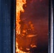 Two firefighters in full protective gear are visible inside a doorway engulfed in intense flames and smoke. The scene appears to be either an active structural fire or a controlled training exercise, with large orange flames climbing the left side of the doorframe. The firefighters are wearing full turnout gear including helmets, SCBA (self-contained breathing apparatus) units, and reflective-striped protective suits.