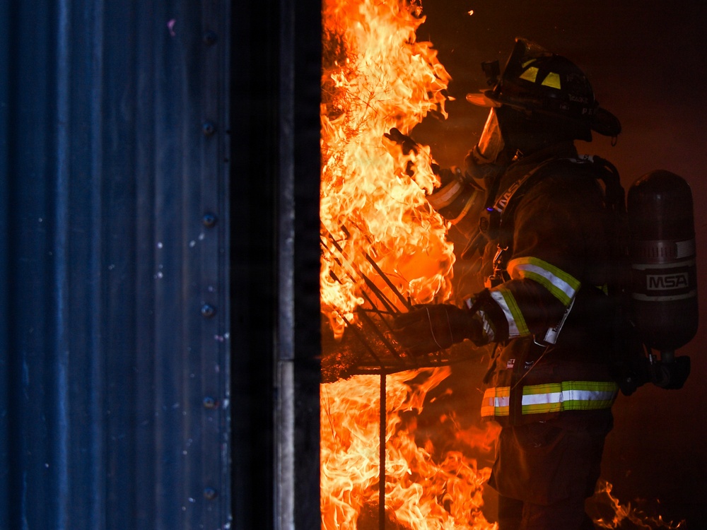 A firefighter in full protective gear is working in close proximity to intense flames bursting through what appears to be a doorway or opening in a structure. The firefighter is equipped with an MSA self-contained breathing apparatus (SCBA) and is wearing standard turnout gear with high-visibility reflective striping. The dramatic lighting from the fire illuminates the scene against a dark background, suggesting nighttime or low-light conditions.