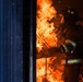 A firefighter in full protective gear is working in close proximity to intense flames bursting through what appears to be a doorway or opening in a structure. The firefighter is equipped with an MSA self-contained breathing apparatus (SCBA) and is wearing standard turnout gear with high-visibility reflective striping. The dramatic lighting from the fire illuminates the scene against a dark background, suggesting nighttime or low-light conditions.