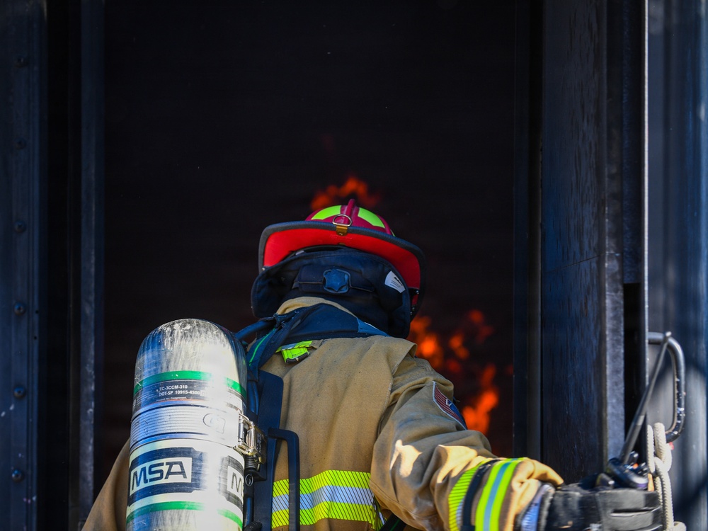 A firefighter in full personal protective equipment is positioned at the entrance of a structure with visible flames and heavy smoke inside. The firefighter is equipped with an MSA self-contained breathing apparatus (SCBA) tank on their back and is wearing a red helmet with a neon yellow trim. The image captures the moment just before or during entry into an active fire environment.