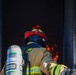A firefighter in full personal protective equipment is positioned at the entrance of a structure with visible flames and heavy smoke inside. The firefighter is equipped with an MSA self-contained breathing apparatus (SCBA) tank on their back and is wearing a red helmet with a neon yellow trim. The image captures the moment just before or during entry into an active fire environment.