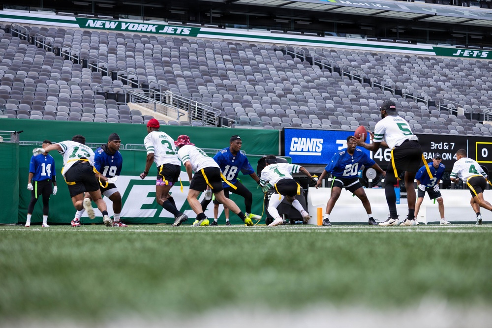 A flag football game is being played on the field at MetLife Stadium, home of the New York Jets and New York Giants. Players wearing white/green and blue uniforms are competing, with a quarterback in white (#5) preparing to throw the ball. The stadium seats are largely empty, suggesting this is a youth or amateur event rather than a professional NFL game.