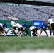 A flag football game is being played on the field at MetLife Stadium, home of the New York Jets and New York Giants. Players wearing white/green and blue uniforms are competing, with a quarterback in white (#5) preparing to throw the ball. The stadium seats are largely empty, suggesting this is a youth or amateur event rather than a professional NFL game.