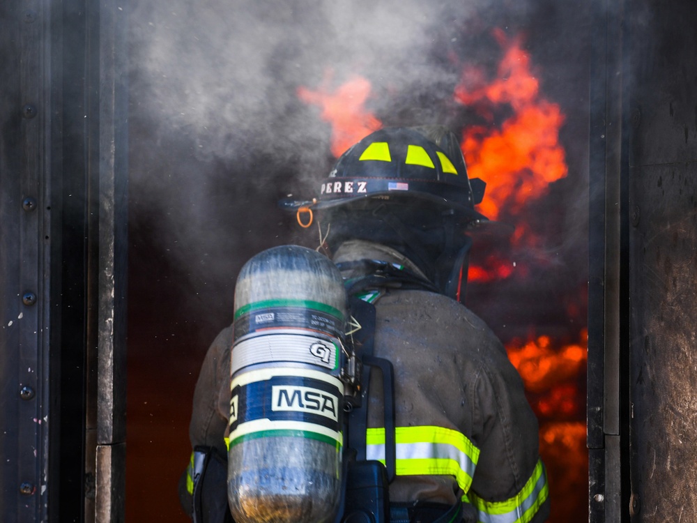 A firefighter identified as 'Perez' is entering or standing at the threshold of a doorway engulfed in flames and smoke. The firefighter is fully equipped with turnout gear, a black helmet with yellow chevron markings, and an MSA self-contained breathing apparatus (SCBA). The image captures an active fire scene with intense orange flames and white smoke visible in the background.