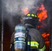 A firefighter identified as 'Perez' is entering or standing at the threshold of a doorway engulfed in flames and smoke. The firefighter is fully equipped with turnout gear, a black helmet with yellow chevron markings, and an MSA self-contained breathing apparatus (SCBA). The image captures an active fire scene with intense orange flames and white smoke visible in the background.