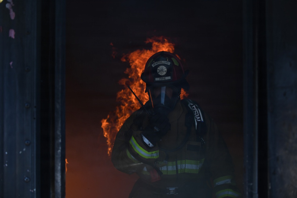 A firefighter in full protective gear stands in a doorway or opening with active flames visible behind them. The firefighter is wearing a helmet with 'FIRE' visible on the front shield, an SCBA (Self-Contained Breathing Apparatus) mask, and a turnout coat with reflective yellow-green striping. The scene is dark and smoke-filled, suggesting an active structural fire response.