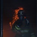 A firefighter in full protective gear stands in a doorway or opening with active flames visible behind them. The firefighter is wearing a helmet with 'FIRE' visible on the front shield, an SCBA (Self-Contained Breathing Apparatus) mask, and a turnout coat with reflective yellow-green striping. The scene is dark and smoke-filled, suggesting an active structural fire response.