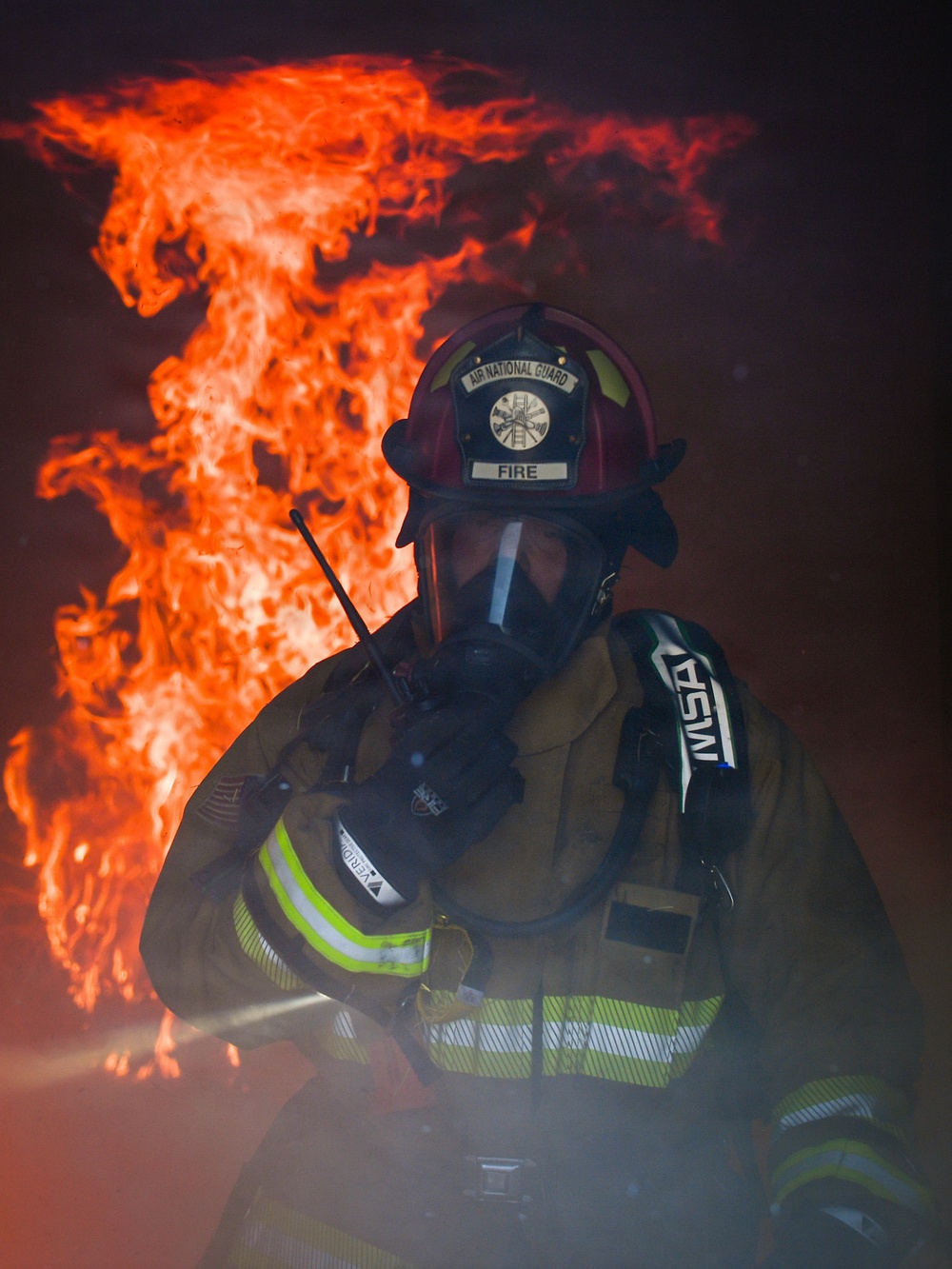 A firefighter affiliated with the Air National Guard is shown in full protective gear against a backdrop of intense flames and smoke. The individual is holding a radio communicator to their face while wearing an MSA self-contained breathing apparatus (SCBA) and a red helmet marked 'FIRE' and 'Air National Guard'. The dramatic composition places the firefighter in the foreground with large orange flames rising behind them in a dark, smoke-filled environment.