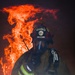 A firefighter affiliated with the Air National Guard is shown in full protective gear against a backdrop of intense flames and smoke. The individual is holding a radio communicator to their face while wearing an MSA self-contained breathing apparatus (SCBA) and a red helmet marked 'FIRE' and 'Air National Guard'. The dramatic composition places the firefighter in the foreground with large orange flames rising behind them in a dark, smoke-filled environment.